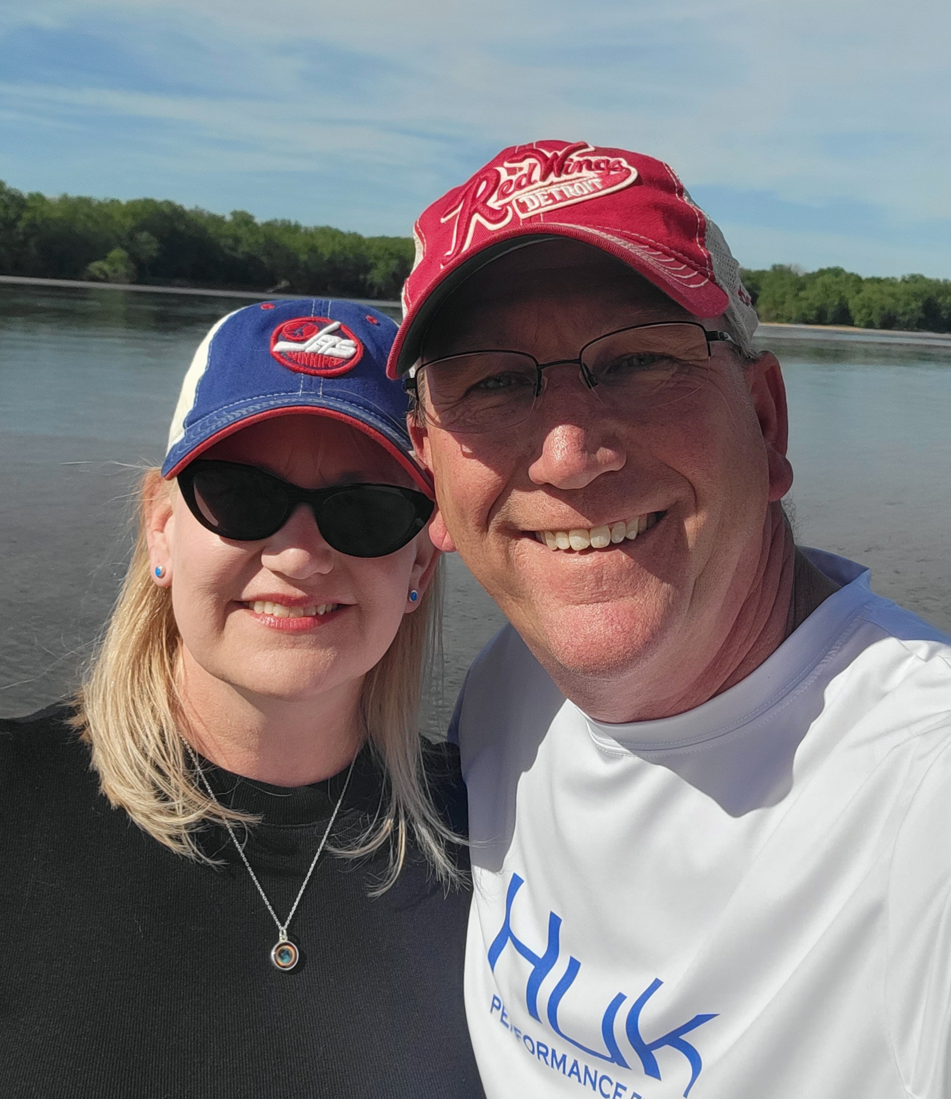 Bradd and Cathie smile. They wear baseball caps and stand in front of a lake.